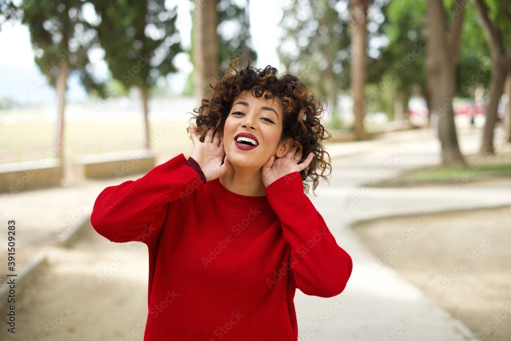 Young beautiful Arab woman standing outdoors wearing red sweater Trying to hear both hands on ear gesture, curious for gossip. Hearing problem, deaf
