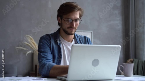 Smiling businessman working on laptop computer at home office. Male professional typing on laptop keyboard at workplace. Portrait of positive business man looking at laptop screen indoors