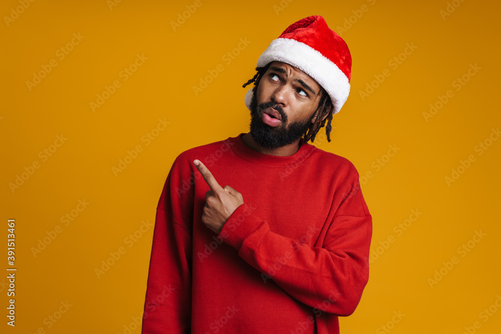 Confused african american guy in Santa Claus hat pointing finger aside Confused african american guy in Santa Claus hat pointing finger aside