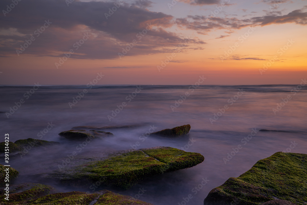 Calm ocean long exposure. Stones covered by green moss in mysterious mist of the sea waves. Concept of nature background. Sunset scenery background. Mengening beach, Bali, Indonesia.