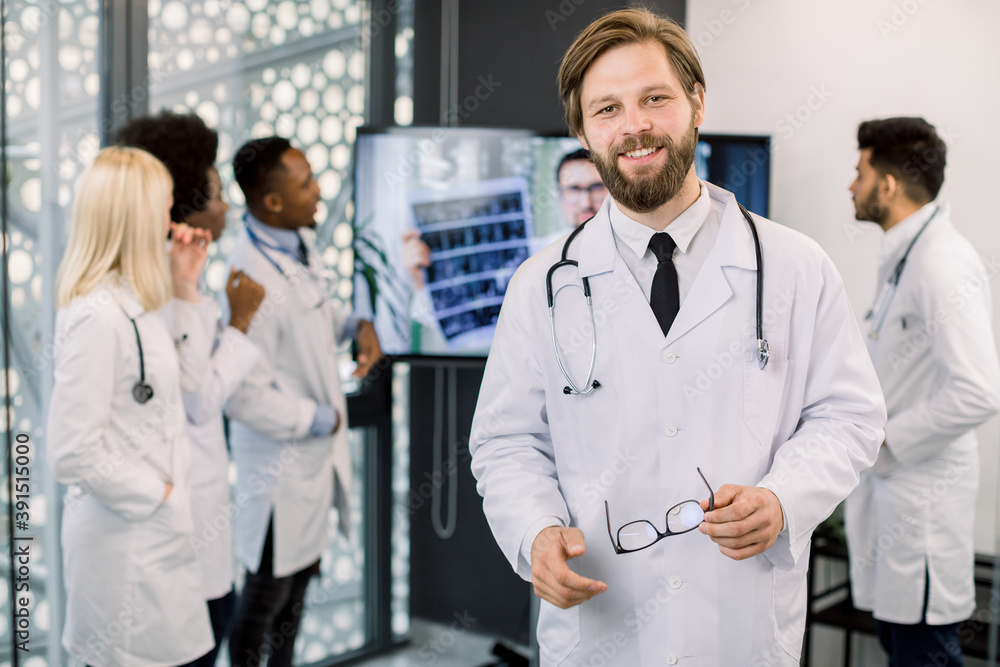 Confident smiling young doctor posing with eyeglasses to camera at the hospital. International medical team working on the background, analyzing patient's CT, consulting with Caucasian doctor online Confident smiling young doctor posing with eyeglasses to camera at the hospital. International medical team working on the background, analyzing patient's CT, consulting with Caucasian doctor online