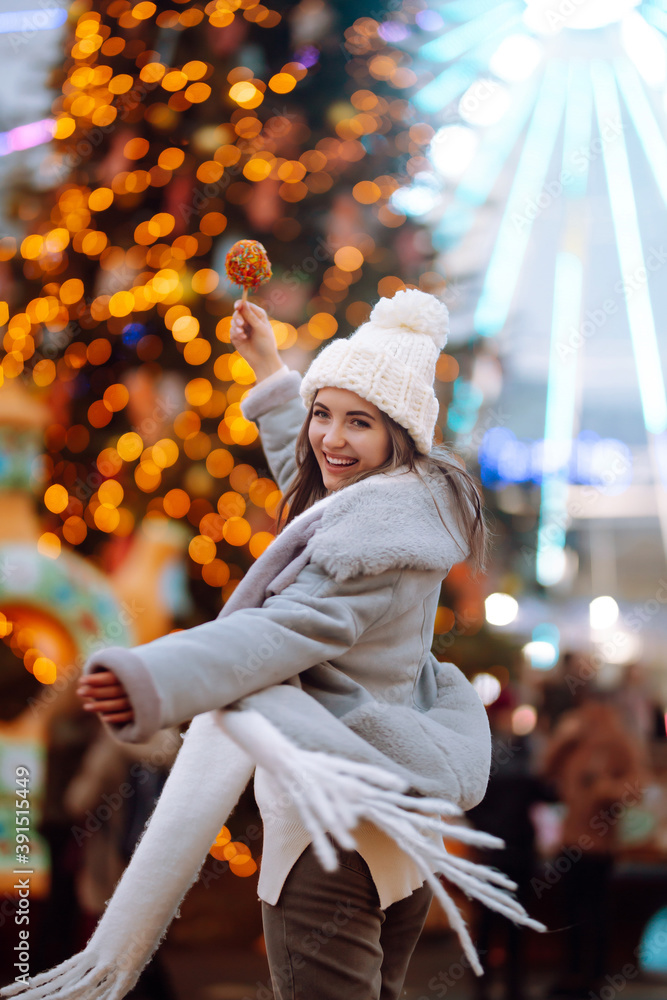 Happy woman on Christmas market with caramelized apple. Outdoor activities on Christmas and New Year. Young woman enjoying winter moments. Lights around.