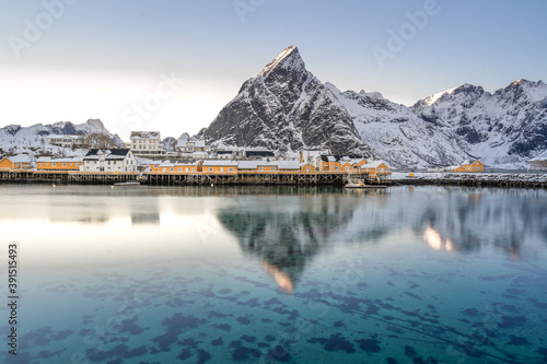 Beautiful winter fishing town on Nordic shore with tall snow mountain backdrop and reflection on the turquoise water. overcast  sky and clouds and evening serene light in cold lonely winter season.  © Jirakit