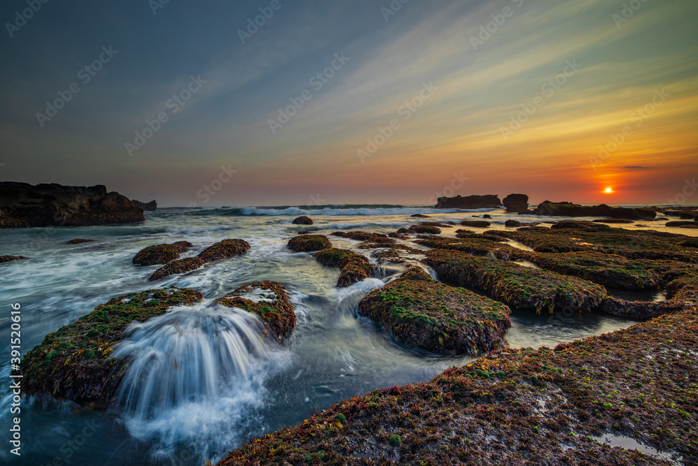 Amazing seascape. Ocean with moving wave. Low tide. Stones covered by green moss and seaweeds. Sun on horizon. Sunset scenery background. Long exposure. Mengening beach, Bali