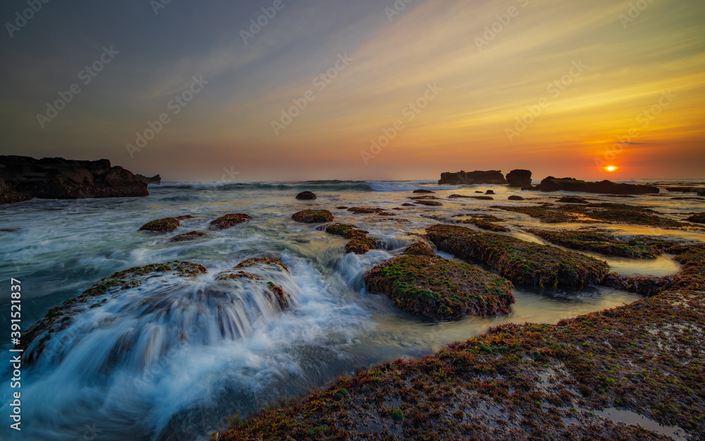 Obraz premium Amazing seascape. Ocean with moving wave. Low tide. Stones covered by green moss and seaweeds. Sun on horizon. Sunset scenery background. Long exposure. Mengening beach, Bali