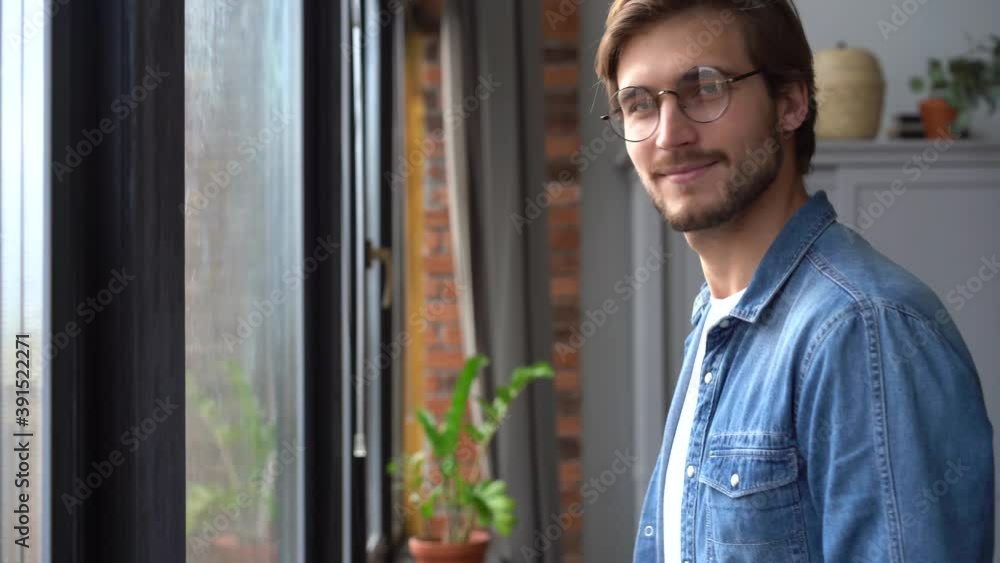 Young man in glasses standing at window at home looking away. Portrait of pensive guy relaxing near window enjoying life Young man in glasses standing at window at home looking away. Portrait of pensive guy relaxing near window enjoying life
