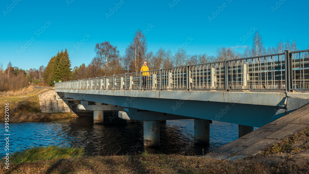 A woman in a yellow jacket walks across a bridge over a river in an autumn afternoon.