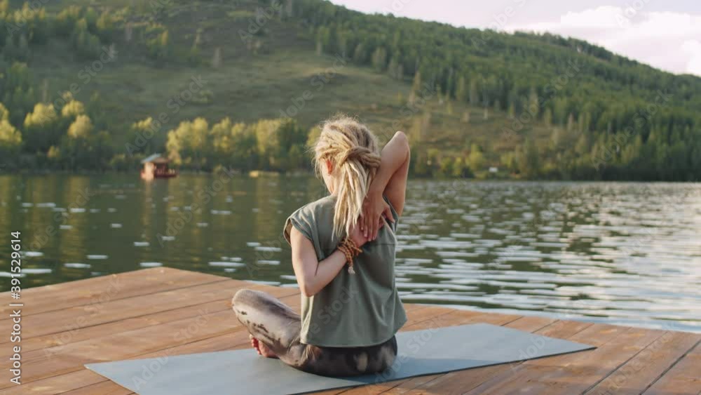 Back view of blonde woman with dreadlocks sitting on mat and holding hands behind back while practicing yoga on wooden lake dock outdoors on summer day Back view of blonde woman with dreadlocks sitting on mat and holding hands behind back while practicing yoga on wooden lake dock outdoors on summer day