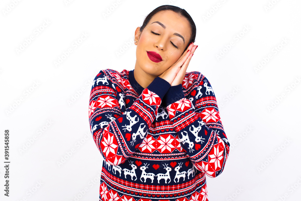 Relax and sleep time. Tired Young beautiful Arab woman wearing Christmas sweater against white wall with closed eyes leaning on palms making sleeping gesture. Relax and sleep time. Tired Young beautiful Arab woman wearing Christmas sweater against white wall with closed eyes leaning on palms making sleeping gesture.