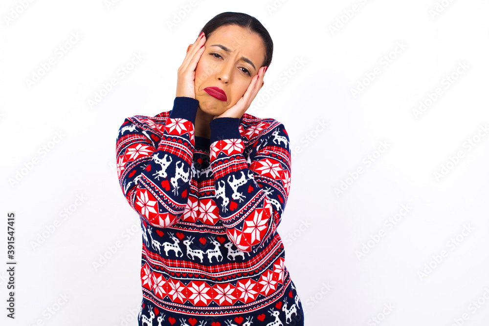 Young beautiful Arab woman wearing Christmas sweater against white wall holding head in hands with unhappy expression watching sad movie about animals and trying not to cry. Young beautiful Arab woman wearing Christmas sweater against white wall holding head in hands with unhappy expression watching sad movie about animals and trying not to cry.