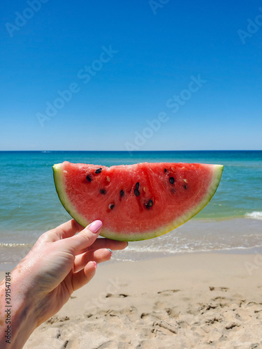 hand holds a slice of watermelon on the beach on a Sunny summer day. Sea and blue sky in the background.