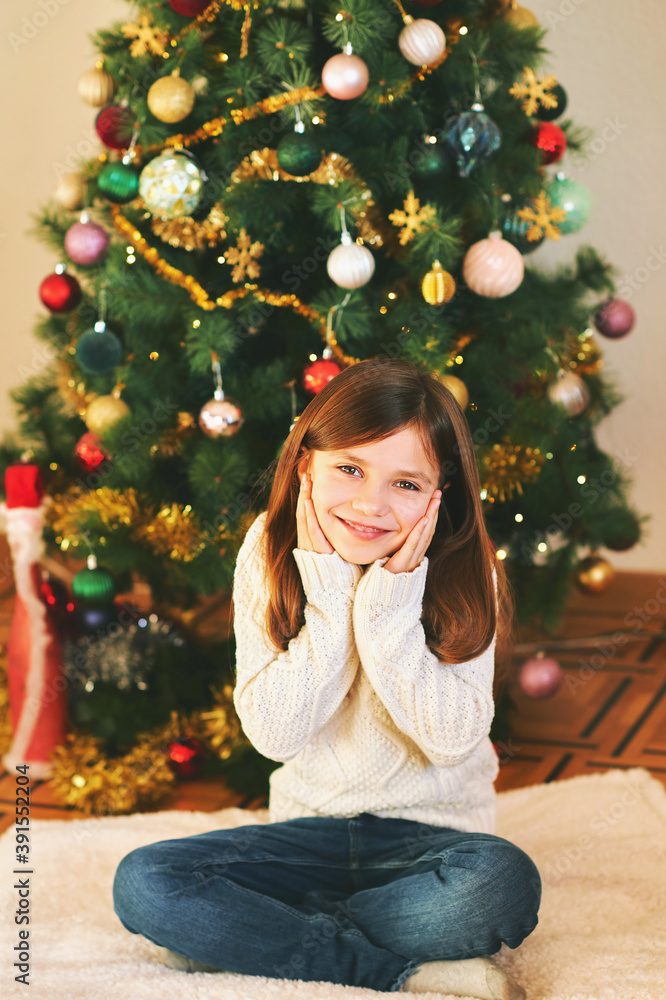 Portrait of sweet little girl sitting next to Christmas tree, wearing white cozy pullover, winter holidays Portrait of sweet little girl sitting next to Christmas tree, wearing white cozy pullover, winter holidays
