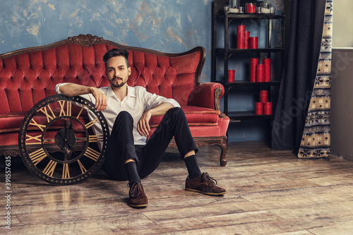 Horizontal portrait of young man sitting in luxury room. Male wearing an expensive suit leaning on a souvenir wooden clock. Professional model posing. Antique style copy space.