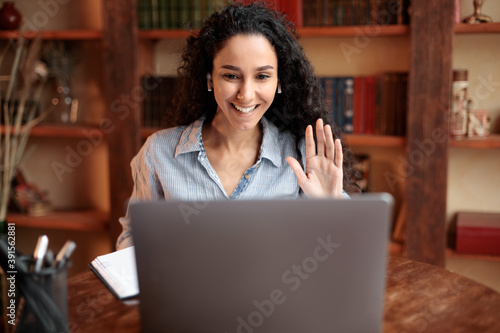 Woman sitting at desk, using computer and waving to webcam