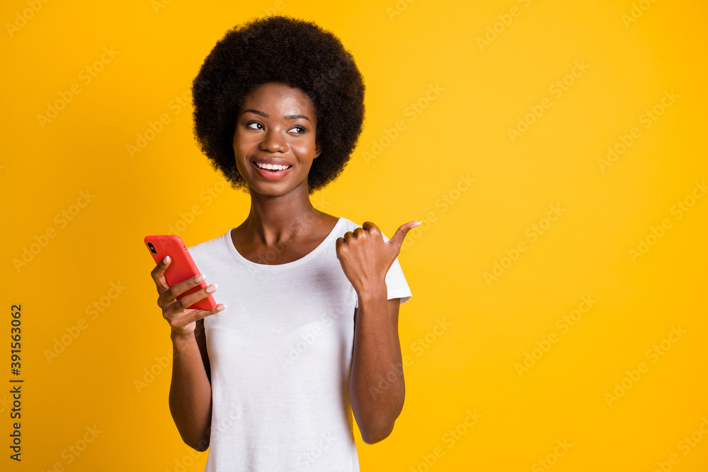 Photo portrait of african american woman pointing at blank space with thumb holding phone in hand wearing white t-shirt isolated on vivid yellow colored background Photo portrait of african american woman pointing at blank space with thumb holding phone in hand wearing white t-shirt isolated on vivid yellow colored background