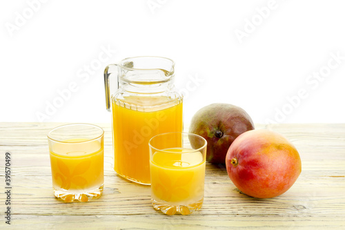 Tasty mango juice in a jug, glasses and ripe mango fruit on a wooden table close-up.