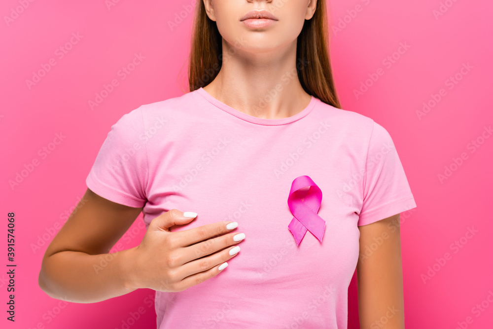 Cropped view of young woman with breast cancer awareness ribbon touching breast on pink background Cropped view of young woman with breast cancer awareness ribbon touching breast on pink background