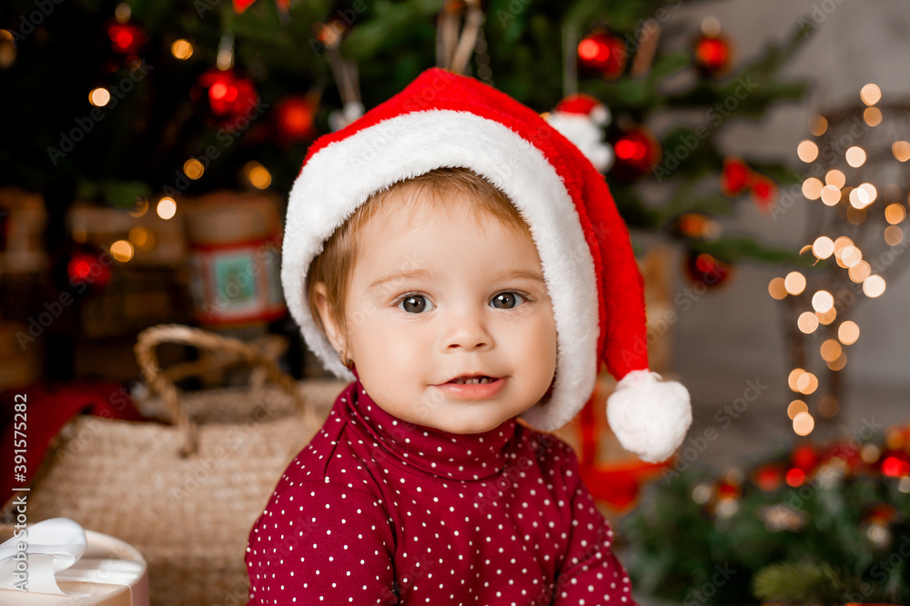 Cute baby Santa sits at home near the Christmas tree with gifts Cute baby Santa sits at home near the Christmas tree with gifts