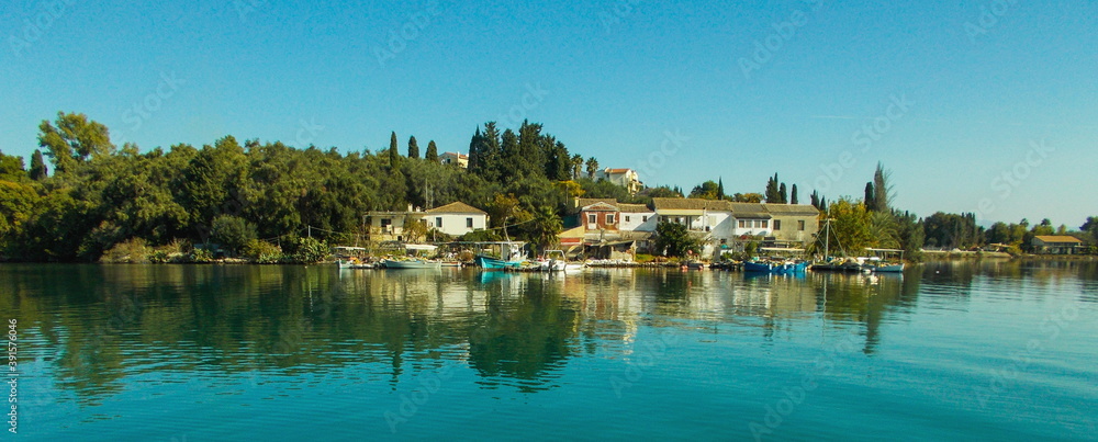 Fototapeta premium panoramic Greek mediteranean fishing village with cottage houses cypress and olive trees and boats on calm sea water