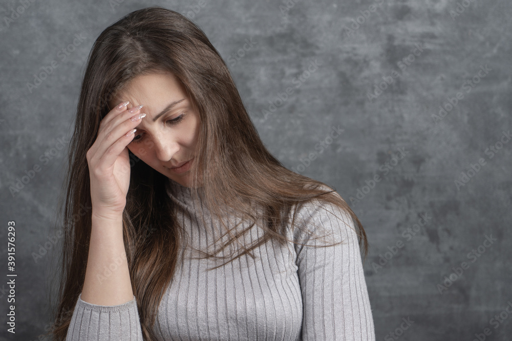 Upset girl with headache, migraine, touches head isolated on gray background. Young brunette woman in stress and crisis with sad face in studio Upset girl with headache, migraine, touches head isolated on gray background. Young brunette woman in stress and crisis with sad face in studio