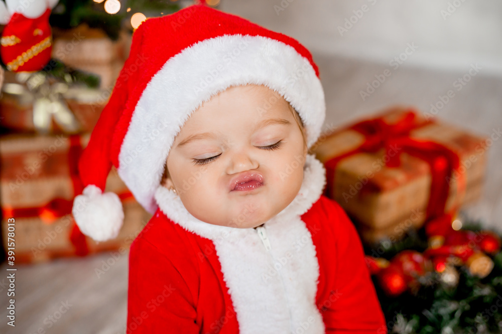 Cute baby Santa sits at home near the Christmas tree with gifts Cute baby Santa sits at home near the Christmas tree with gifts