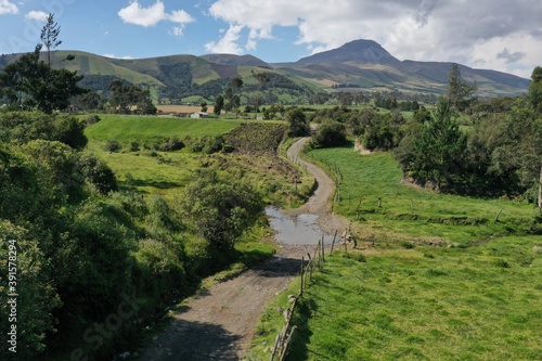 A small dirtroad that is running through two meadows with lush green grass separeted by a fence of wooden pools covered by a puddle with mountains in the background photo