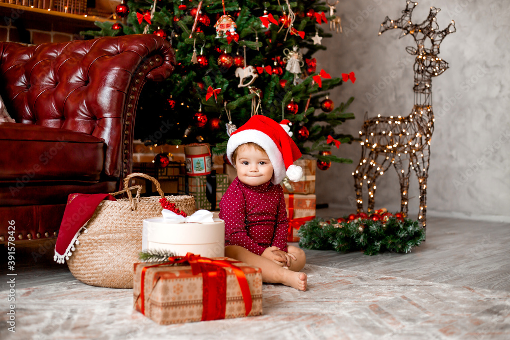 Cute baby Santa sits at home near the Christmas tree with gifts Cute baby Santa sits at home near the Christmas tree with gifts