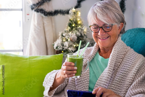 Smiling senior woman sitting at home and holding a glass of green smoothie while looking at her smart phone. Healthy detox vegan diet with fruits and vegetables. Christmas decoration on the background