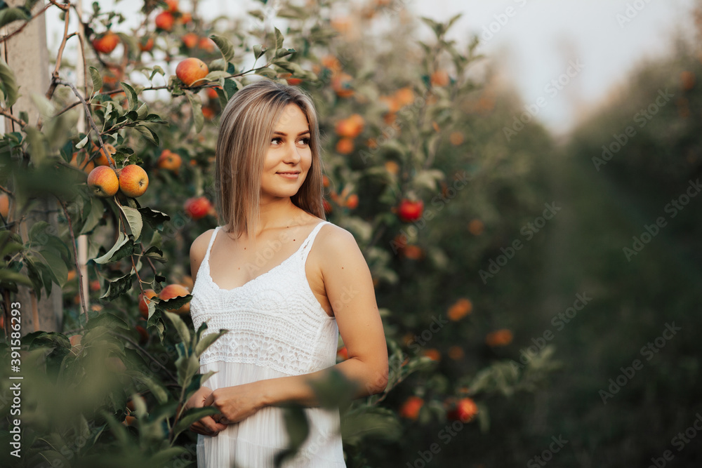 Pretty blonde girl in white dress posing in the apple orchard, smiling and looking away.