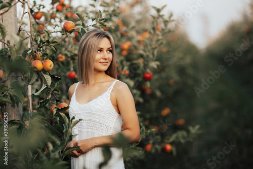 Obraz na płótnie Pretty blonde girl in white dress posing in the apple orchard, smiling and looking away. w salonie
