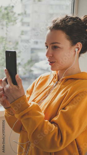 Young brunette woman in headphones and yellow hoodie holding smartphone for video online calling, making selfie using internet connection, sharing data on social media,standing near window. Vertical