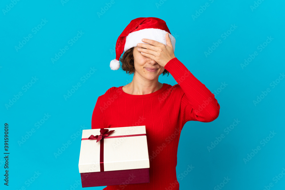 English girl with christmas hat holding a present isolated on blue background covering eyes by hands. Do not want to see something English girl with christmas hat holding a present isolated on blue background covering eyes by hands. Do not want to see something