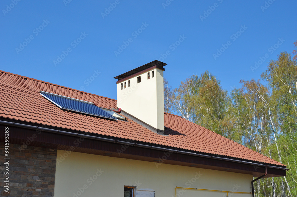 A close-up of an energy efficient detached house's clay roof with a solar water heating thermal panel, a skylight, a chimney and a rain gutter system against blue sky.