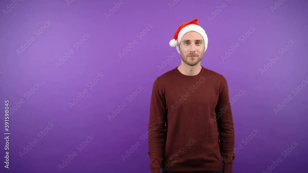funny young attractive bearded man in a christmas santa hat points his finger next to the blank space. man over isolated purple background. 4K funny young attractive bearded man in a christmas santa hat points his finger next to the blank space. man over isolated purple background. 4K