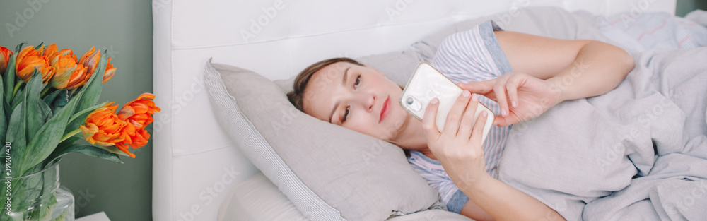 Middle age woman lying in bed with smartphone surfing Internet. Young woman talking with friend in social media. Digital online Internet addiction. Morning routine. Web banner header. Middle age woman lying in bed with smartphone surfing Internet. Young woman talking with friend in social media. Digital online Internet addiction. Morning routine. Web banner header.