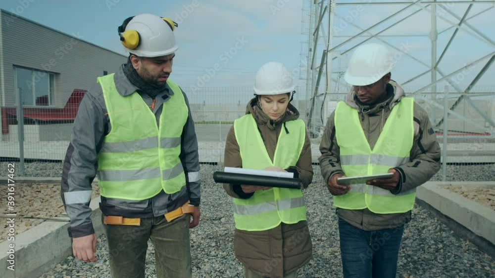 Multinational electric power station engineers in protective helmets and vests hold folders and discuss work on windy autumn day Multinational electric power station engineers in protective helmets and vests hold folders and discuss work on windy autumn day