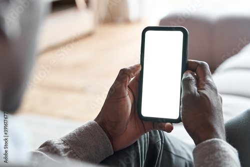 African american man holding smart phone with mockup white blank display, empty screen for app ads sitting on couch at home. Mobile applications technology concept, over shoulder close up view.