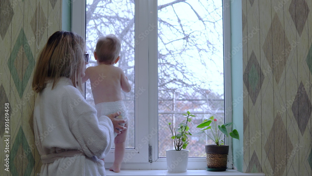 Disheveled mom in a dressing gown stands with a child in front of the window Disheveled mom in a dressing gown stands with a child in front of the window