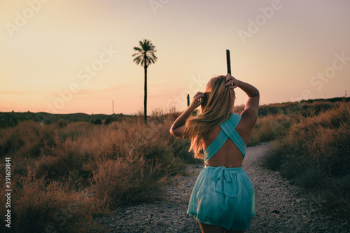 Girl back with blonde hair wearing a teal colour dress and walking through a path with a palmtree in a natural background at sunset