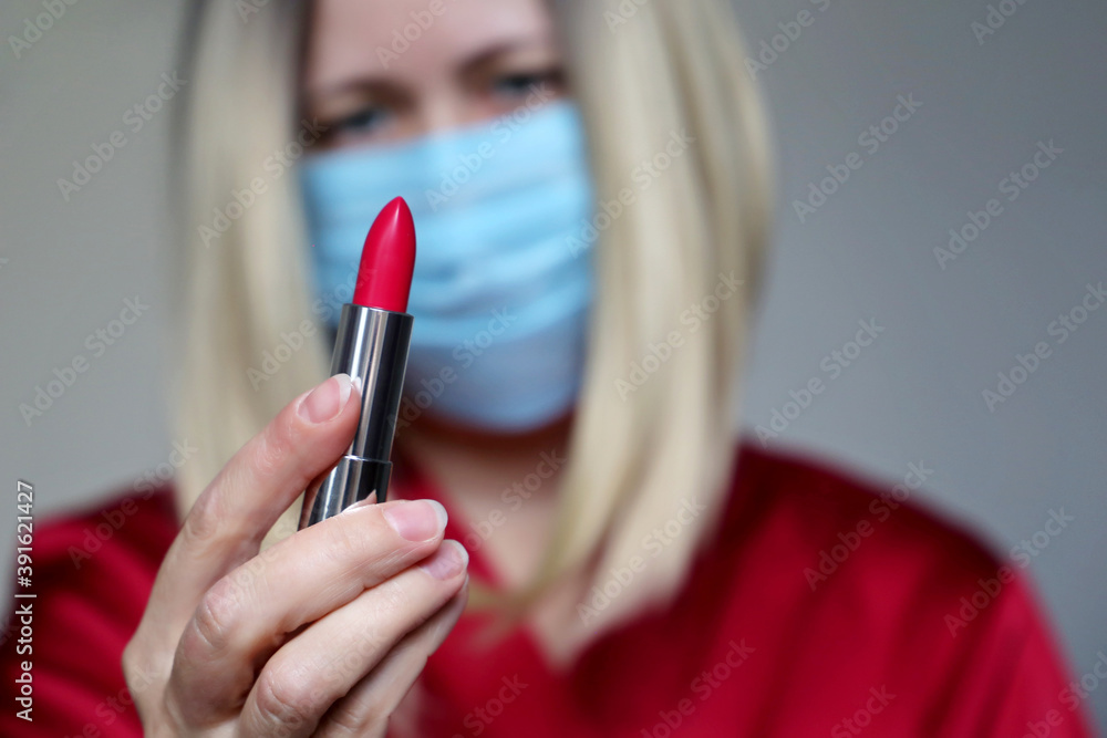 Woman in disposable medical mask looking on red lipstick in her hand. Makeup and cosmetics during the coronavirus pandemic Woman in disposable medical mask looking on red lipstick in her hand. Makeup and cosmetics during the coronavirus pandemic