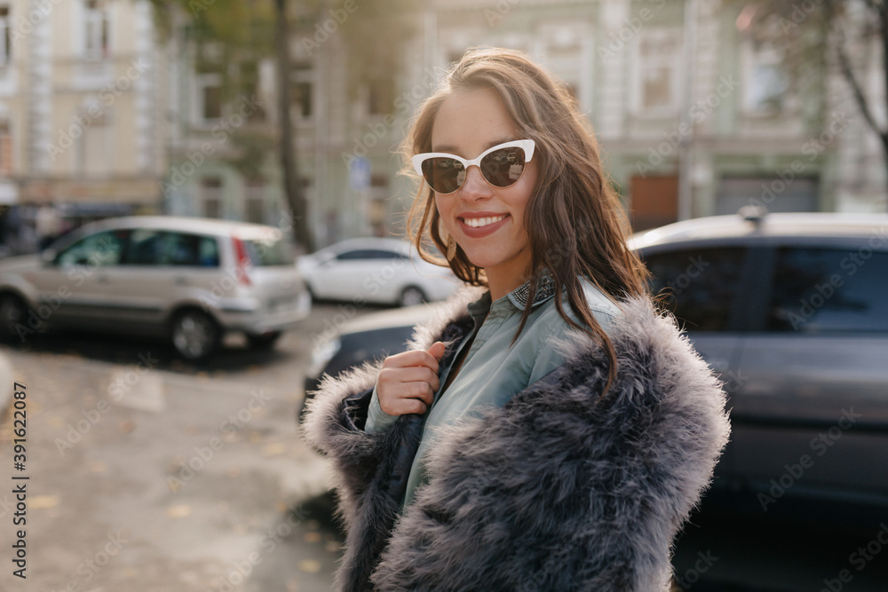 Emotional trendy model in fur coat posing on background of street. Portrait of graceful european girl with long curly hairstyle having fun during outdoor photoshoot. Emotional trendy model in fur coat posing on background of street. Portrait of graceful european girl with long curly hairstyle having fun during outdoor photoshoot.