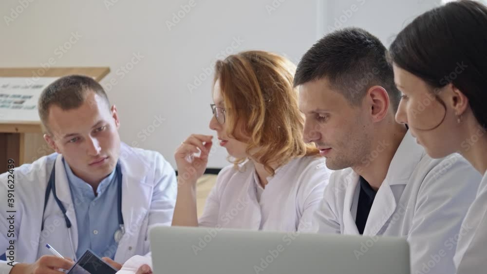 Team of Four Doctors in White Coats Sitting at Table in Hospital and Working With a Laptop Discussing Symptoms of Disease for Further Treatment.Practical Work of Interns.Medical Examination. Team of Four Doctors in White Coats Sitting at Table in Hospital and Working With a Laptop Discussing Symptoms of Disease for Further Treatment.Practical Work of Interns.Medical Examination.