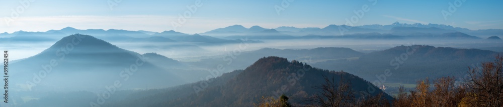 panorama of a mystical and romantic mountain scenery with layers of hilltops softly peaking out of white clouds. Fog flowing through the valleys with alps in the background. 