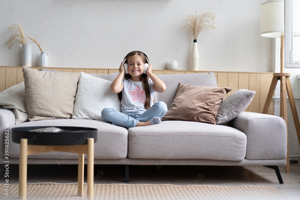 Adorable small schoolgirl sitting on sofa, wearing modern wireless headphones, listening to favorite music, Cute little child resting on couch Adorable small schoolgirl sitting on sofa, wearing modern wireless headphones, listening to favorite music, Cute little child resting on couch