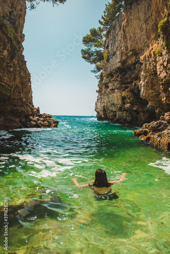 view from behind young pretty woman swimming in sea with clear azure water between rocks