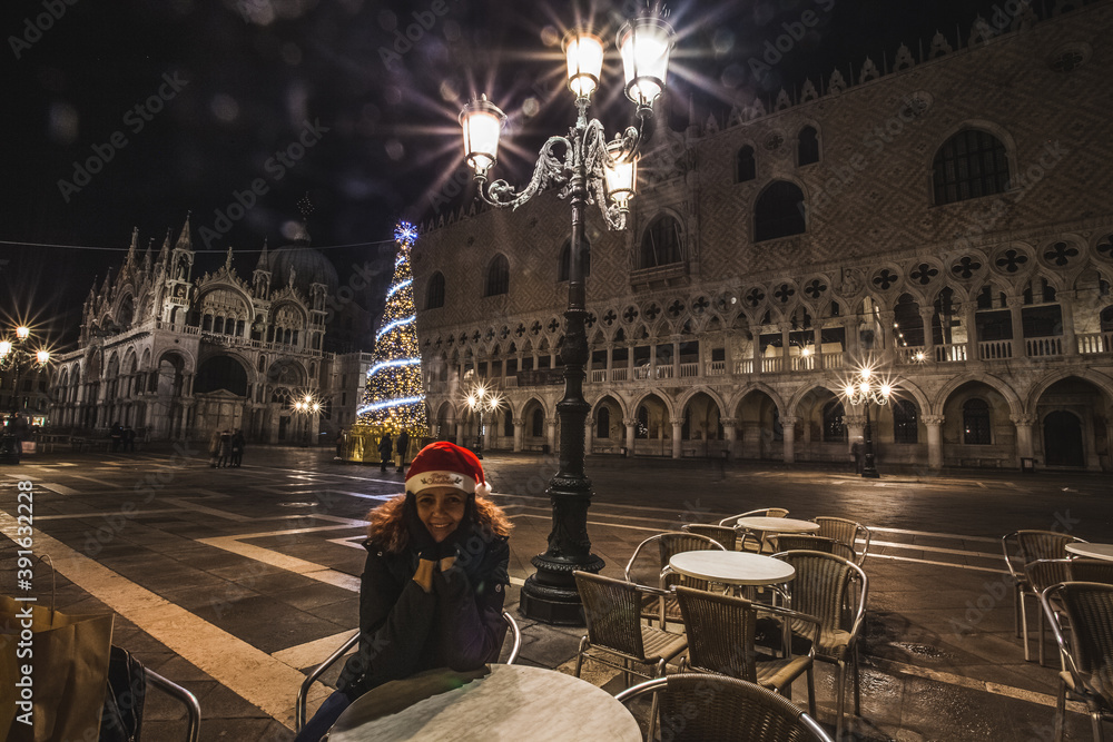 Brazilian woman with Christmas cap sitting at a table in a bar in Piazza San Marco in the evening and in a Christmas context, Venice, Italy
