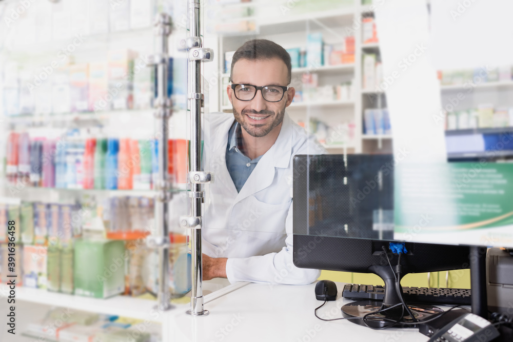 happy pharmacist in white coat and eyeglasses looking at camera near computer monitor in drugstore