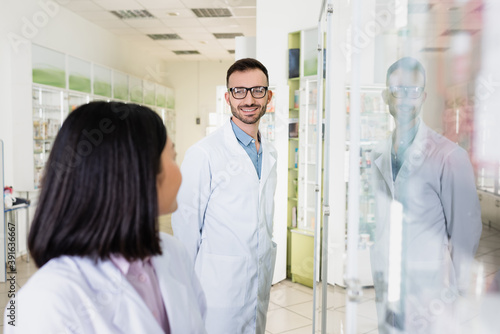 cheerful pharmacist in eyeglasses and white coat looking at brunette asian colleague on blurred foreground