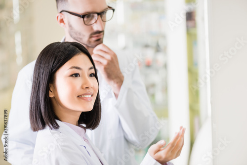 smiling asian pharmacist in white coat pointing with hand near colleague in eyeglasses on blurred background