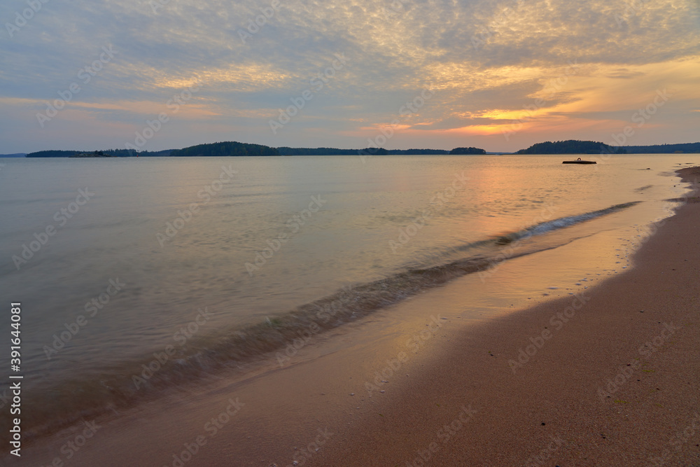 Sunset on the sandy beach with soft waves hitting sand and cloudy sky on the background.